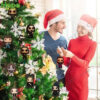 A smiling man and woman wearing Santa hats are decorating a Christmas tree with Ozzy Osbourne ornaments!